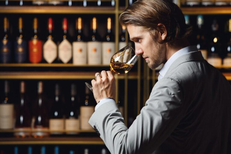 Sommelier appréciant un vin blanc dans un verre en le sentant, debout dans une cave contre une étagère pleine.